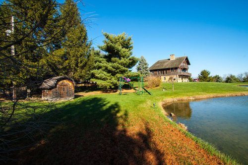 A scenic view of a house by a pond, surrounded by trees and a small shed, under a clear blue sky.