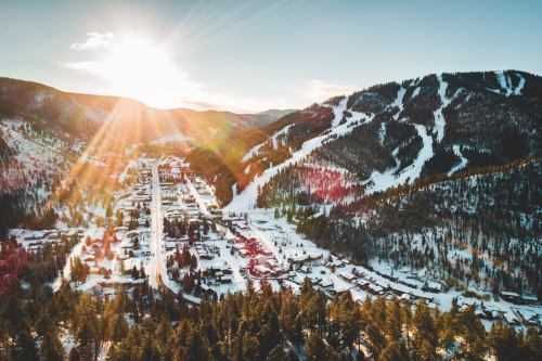 Aerial view of a snowy mountain town with ski slopes, surrounded by trees and bathed in warm sunlight.