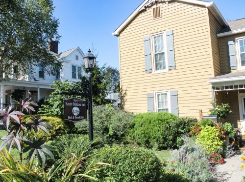 A charming yellow inn with shutters, surrounded by lush greenery and colorful flowers, featuring a sign for Scott Station Inn.