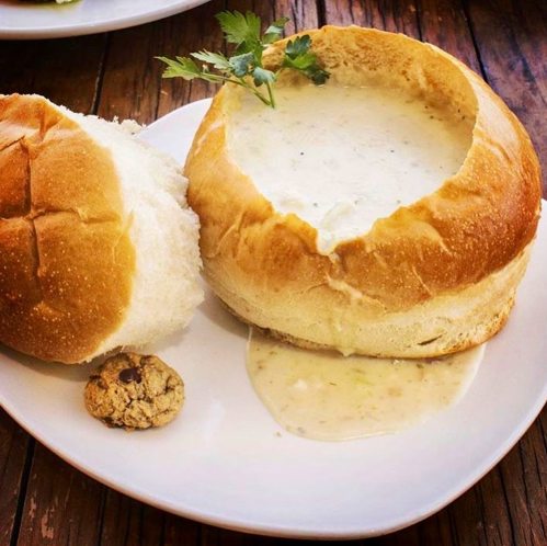 A bread bowl filled with creamy soup, garnished with a sprig of cilantro, alongside a small cookie on a plate.