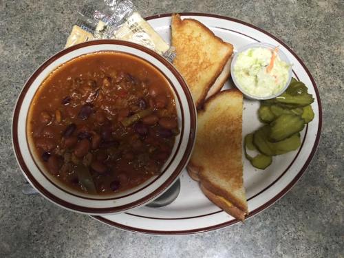 A bowl of chili with beans, toast, coleslaw, pickles, and crackers on a plate.
