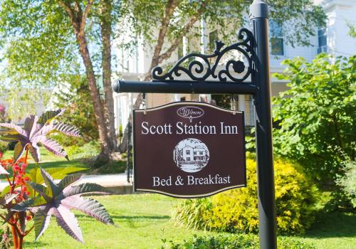 Sign for Scott Station Inn Bed & Breakfast, surrounded by greenery and flowers in a sunny outdoor setting.