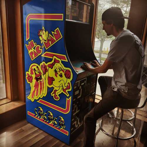 A person playing Ms. Pac-Man at an arcade cabinet in a cozy setting with wooden decor.