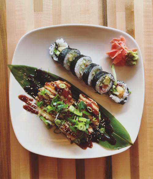 A plate of sushi featuring rolls with seaweed, rice, and toppings, alongside a grilled dish drizzled with sauce and garnished.