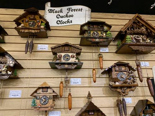 A display of Black Forest cuckoo clocks on a wooden wall, featuring various designs and prices.