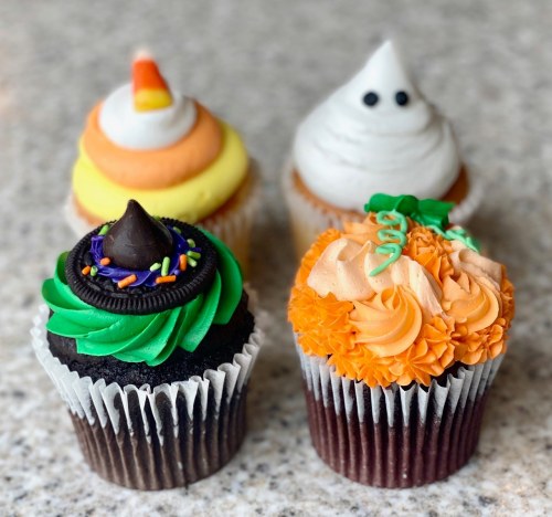 Four festive cupcakes: a witch hat, pumpkin, ghost, and candy corn design, all decorated with vibrant frosting.