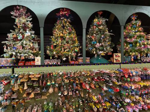 Three decorated Christmas trees with lights and ornaments, displayed above a wall of colorful holiday ornaments.