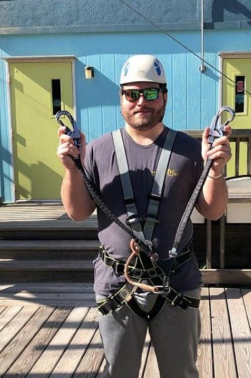 A man wearing a helmet and sunglasses holds safety harness straps, standing on a wooden deck with colorful buildings behind him.