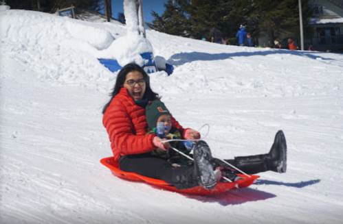 A woman and a child joyfully sledding down a snowy hill on a bright, sunny day.