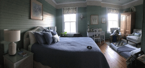 Cozy bedroom with green walls, a large bed, a chair, and a wooden wardrobe, featuring natural light and decorative plants.