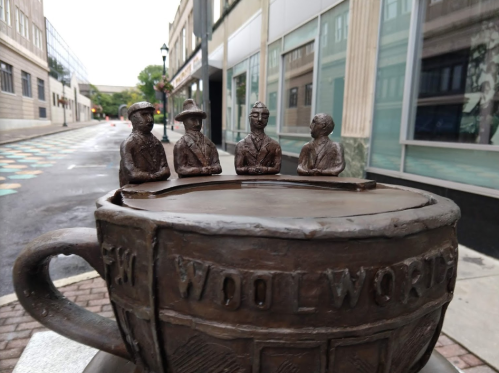 A bronze sculpture of four figures seated around a large cup, set on a city street with buildings in the background.