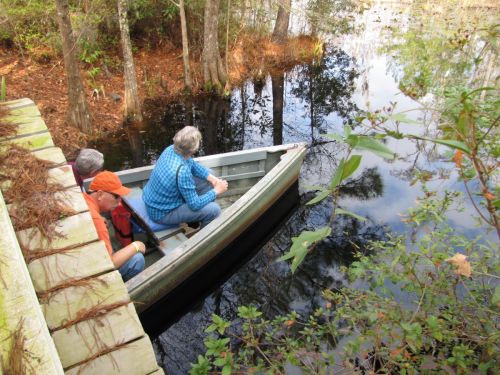 Two people sit in a small boat on a calm, reflective waterway surrounded by trees and foliage.