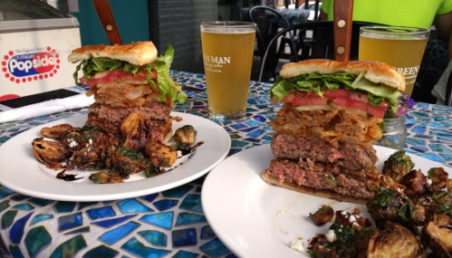 A towering burger with lettuce, tomato, and crispy onions, served with roasted Brussels sprouts and two beers on a colorful table.