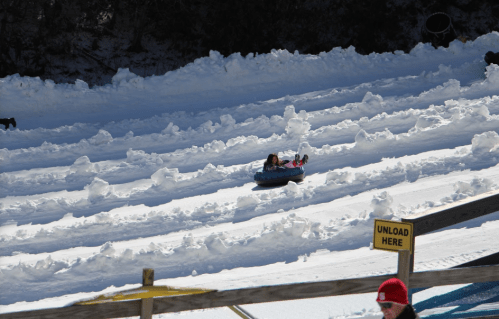 A child tubing down a snowy slope, with tracks in the snow and a person in a red hat nearby.