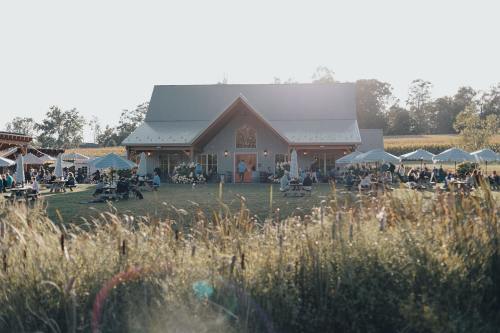 A rustic barn venue surrounded by people enjoying an outdoor gathering, with picnic tables and umbrellas in a grassy field.