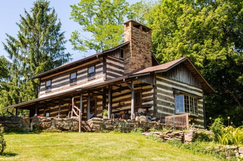 A rustic log cabin with a stone chimney, surrounded by greenery and a well-maintained lawn.