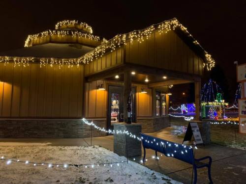 A warmly lit building adorned with holiday lights, surrounded by festive decorations and a snowy pathway.