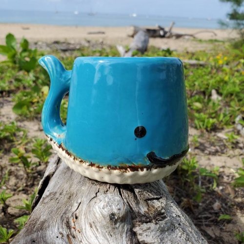 A whimsical blue whale-shaped mug sits on a log by the beach, surrounded by greenery and a serene ocean view.