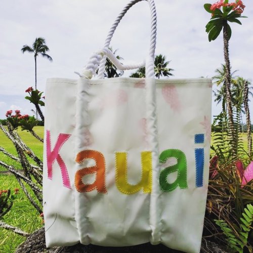 A white tote bag with "Kauai" in colorful letters, set against a tropical background with palm trees and flowers.
