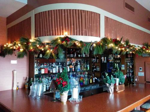 A festive bar decorated with greenery and lights, showcasing various bottles and drinks on the shelves behind.