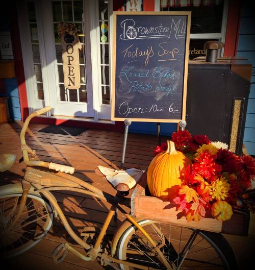 A vintage bicycle with a wooden basket filled with flowers and a pumpkin, next to a sign for today's soup at Brownstone Mill.