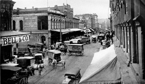 Historic street scene with horse-drawn carriages, early 20th-century buildings, and bustling activity.