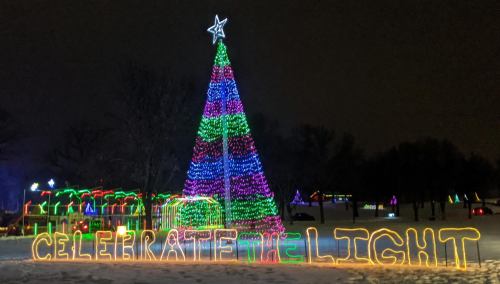 Colorful holiday lights form a large Christmas tree and the words "CELEBRATE the LIGHT" in a snowy park at night.