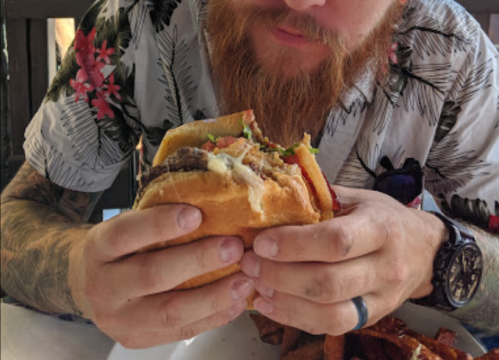 A person with a beard holds a large burger, wearing a floral shirt, with sweet potato fries on the table.