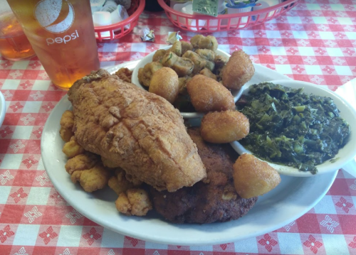 A plate of fried fish, hushpuppies, okra, collard greens, and a drink on a checkered tablecloth.