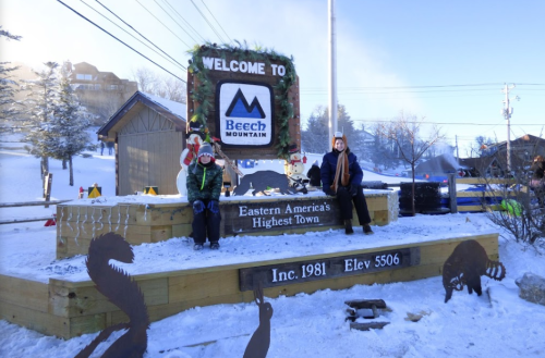 Two people sit on a wooden platform with a sign that reads "Welcome to Beech Mountain" in a snowy landscape.