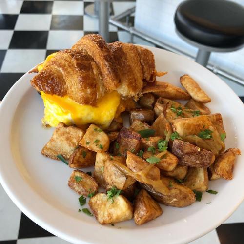 A plate with a cheesy croissant sandwich and a side of golden, diced, and seasoned potatoes.