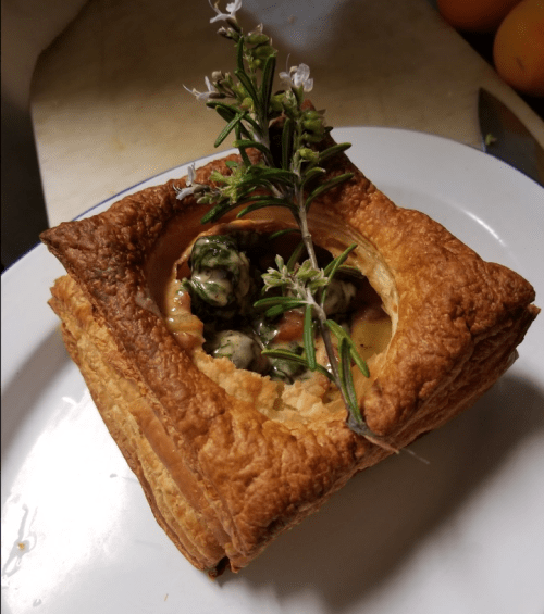 A flaky pastry filled with greens and topped with a sprig of rosemary, served on a white plate.
