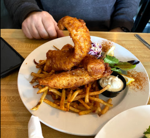 A plate of crispy fried fish fillets on a bed of fries, served with coleslaw and a small bowl of dipping sauce.