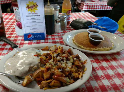 A plate of breakfast food with hash browns, chicken, and gravy, alongside a pancake with syrup, on a checkered tablecloth.