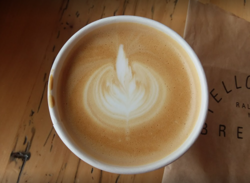 A close-up of a latte with a leaf-shaped foam design, placed on a wooden table next to a paper bag.
