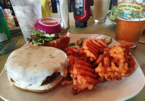 A plate featuring a cheeseburger with lettuce, tomato, and onion, alongside sweet potato waffle fries and a beer.