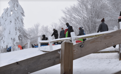 A snowy hill with people sledding down, surrounded by trees and a wooden railing in the foreground.