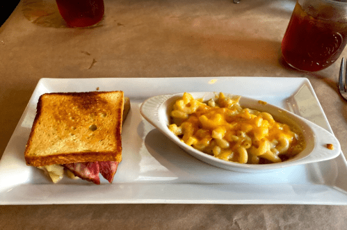 A plate with a toasted sandwich and a side of creamy macaroni and cheese. Drinks are visible in the background.