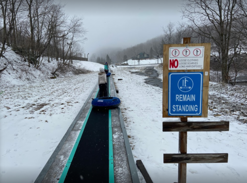 A snowy landscape with a sign that says "REMAIN STANDING" next to a conveyor belt for tubing.