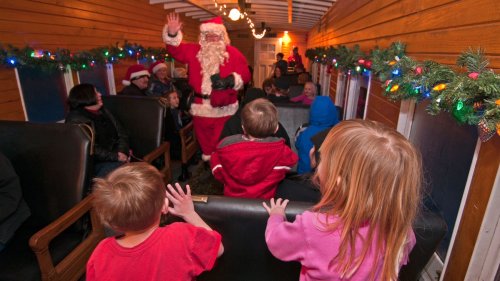 Children watch as Santa waves from the front of a decorated train car filled with festive lights and holiday cheer.
