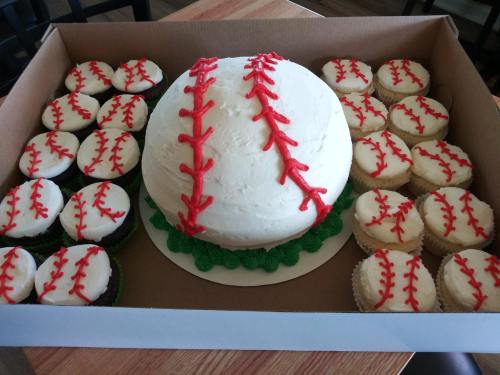 A large baseball-shaped cake surrounded by cupcakes decorated like baseballs, all in a cardboard box.