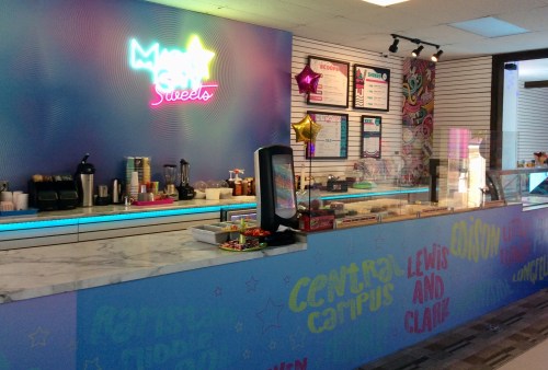 Brightly colored dessert shop counter with neon sign, menu boards, and a variety of sweets displayed.