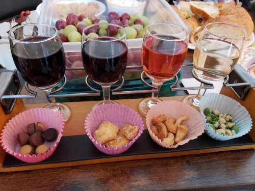 A wooden tray holds four glasses of wine and five cups of snacks, including chocolates, pastries, and nuts.