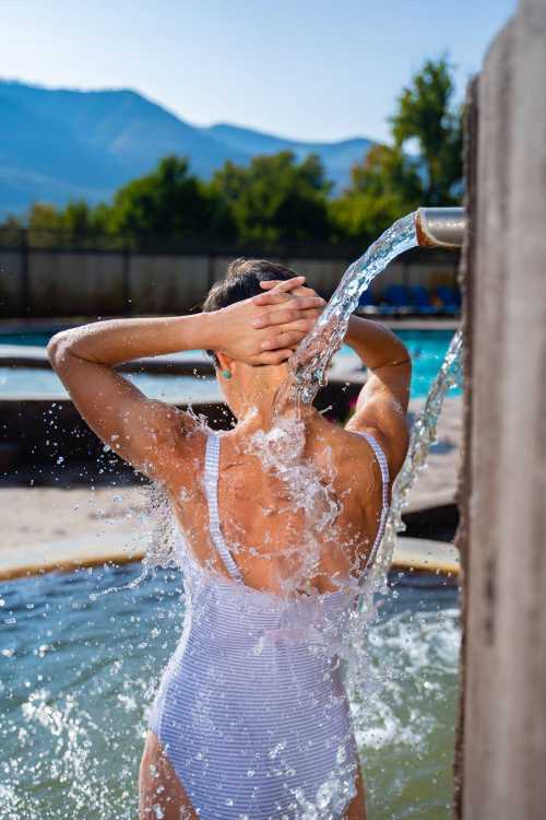 A person in a white swimsuit stands under a water spout, with water splashing around them and mountains in the background.