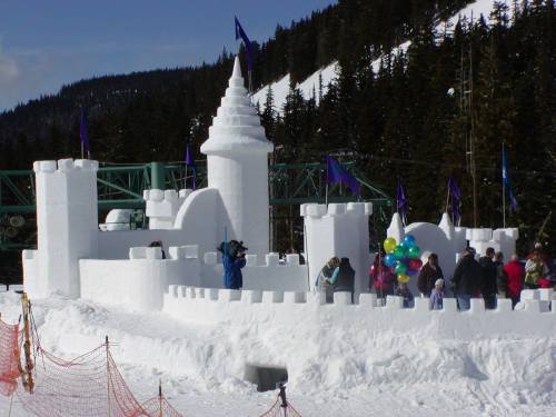A large snow castle with towers and flags, surrounded by people holding balloons, set against a snowy mountain backdrop.