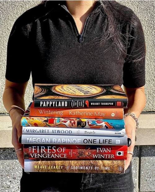 A person holds a stack of books, including titles by Megan Rapinoe and Margaret Atwood, against a textured background.