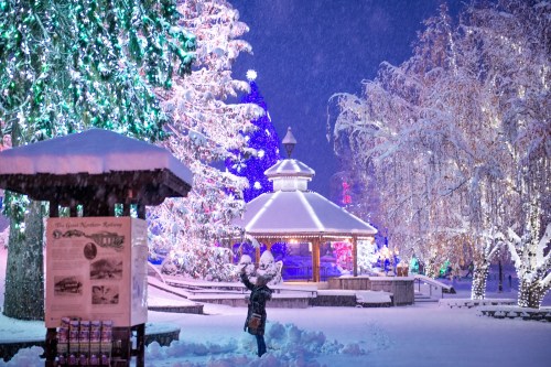 A snowy park scene with colorful holiday lights, a gazebo, and a person playing in the snow under falling flakes.