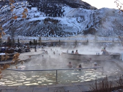 A steaming hot spring surrounded by snow-covered mountains, with people relaxing in the water.
