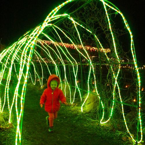 A child in a red coat runs through a glowing green light tunnel at night, surrounded by festive decorations.