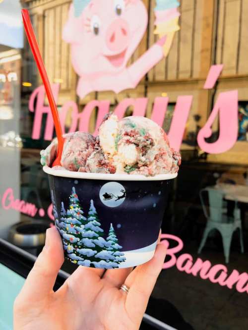 A hand holding a cup of colorful ice cream in front of a shop with a pink pig logo and festive decorations.